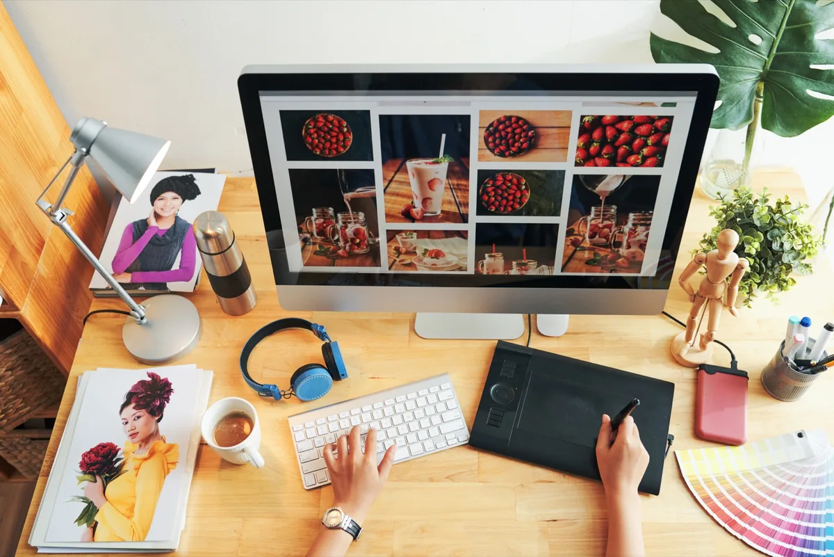 Schreibtisch mit Bildschirm voller Food-Fotos, Tastatur, Grafiktablett, Kopfhörern und aufgeschlagenem Magazin