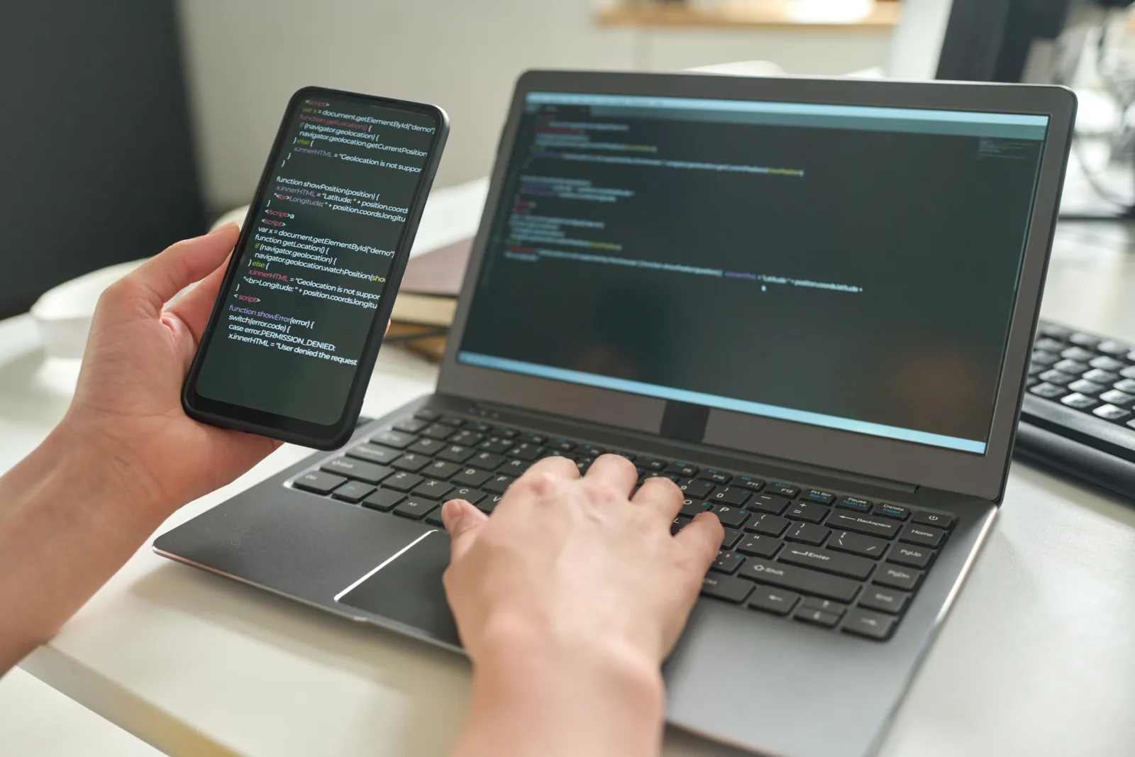 Hände an einem Notebook mit Programmcode, in der anderen Hand ein Smartphone, das ebenfalls Code zeigt