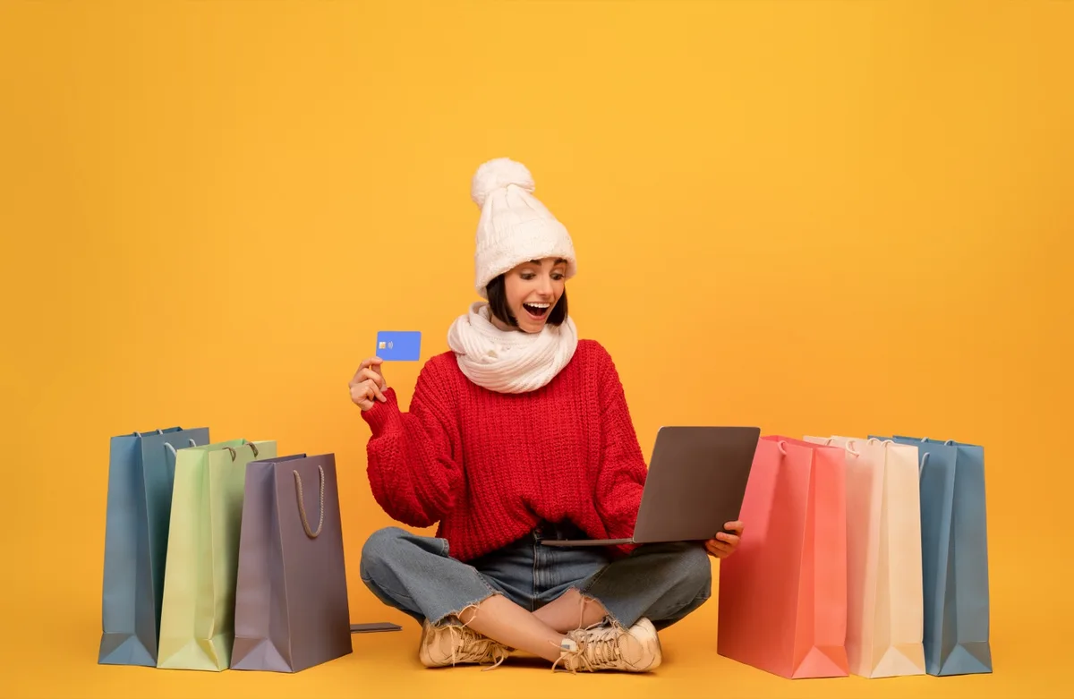 Eine Frau mit Mütze sitzt zwischen Einkaufstüten, hält eine Kreditkarte und einen Laptop
