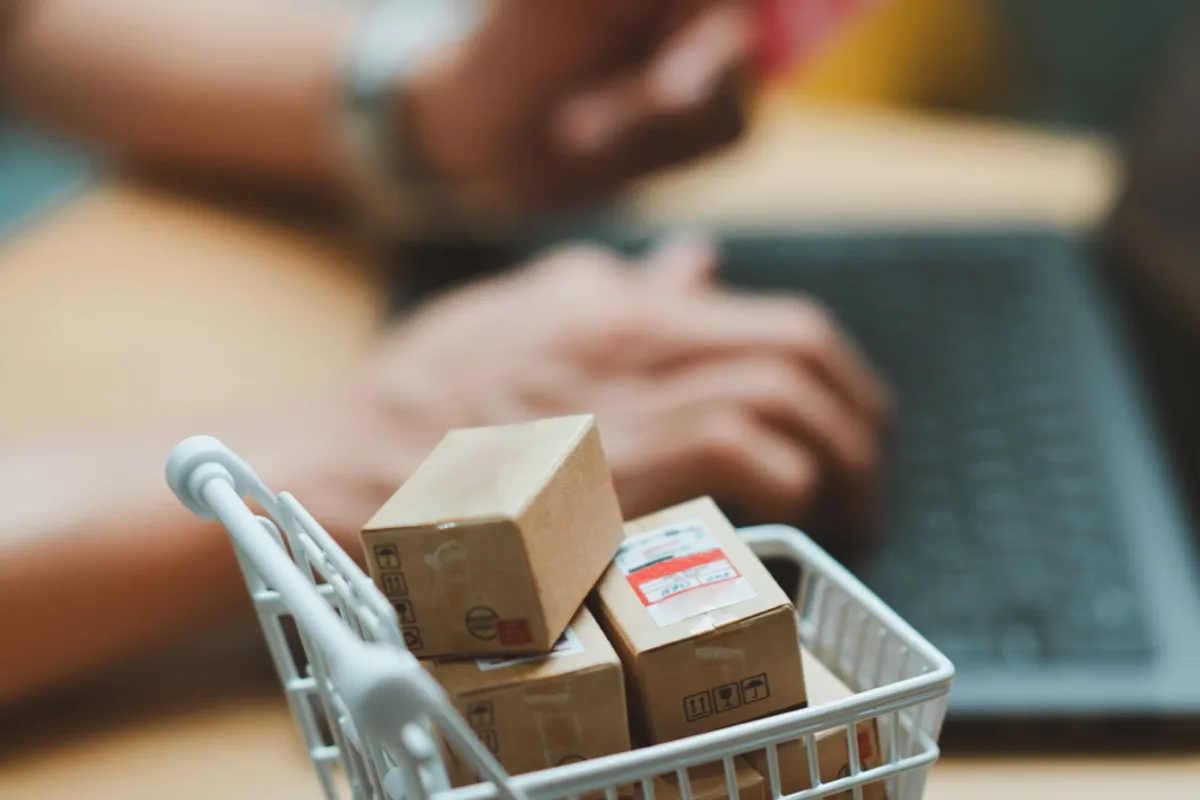 Ein kleiner Einkaufswagen mit Paketen, dahinter unscharf Hände an einem Laptop