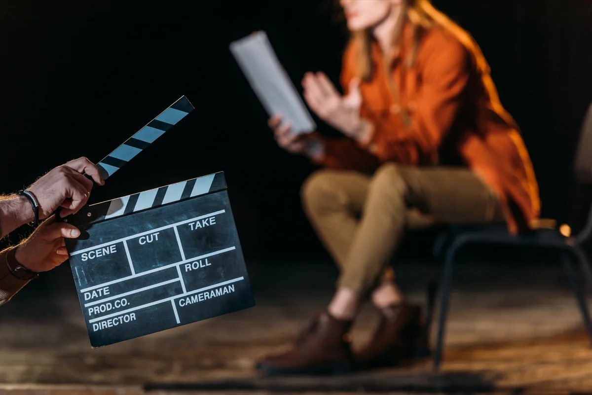Eine Hand hält eine Filmklappe, dahinter unscharf eine sitzende Person mit einem Blatt Papier