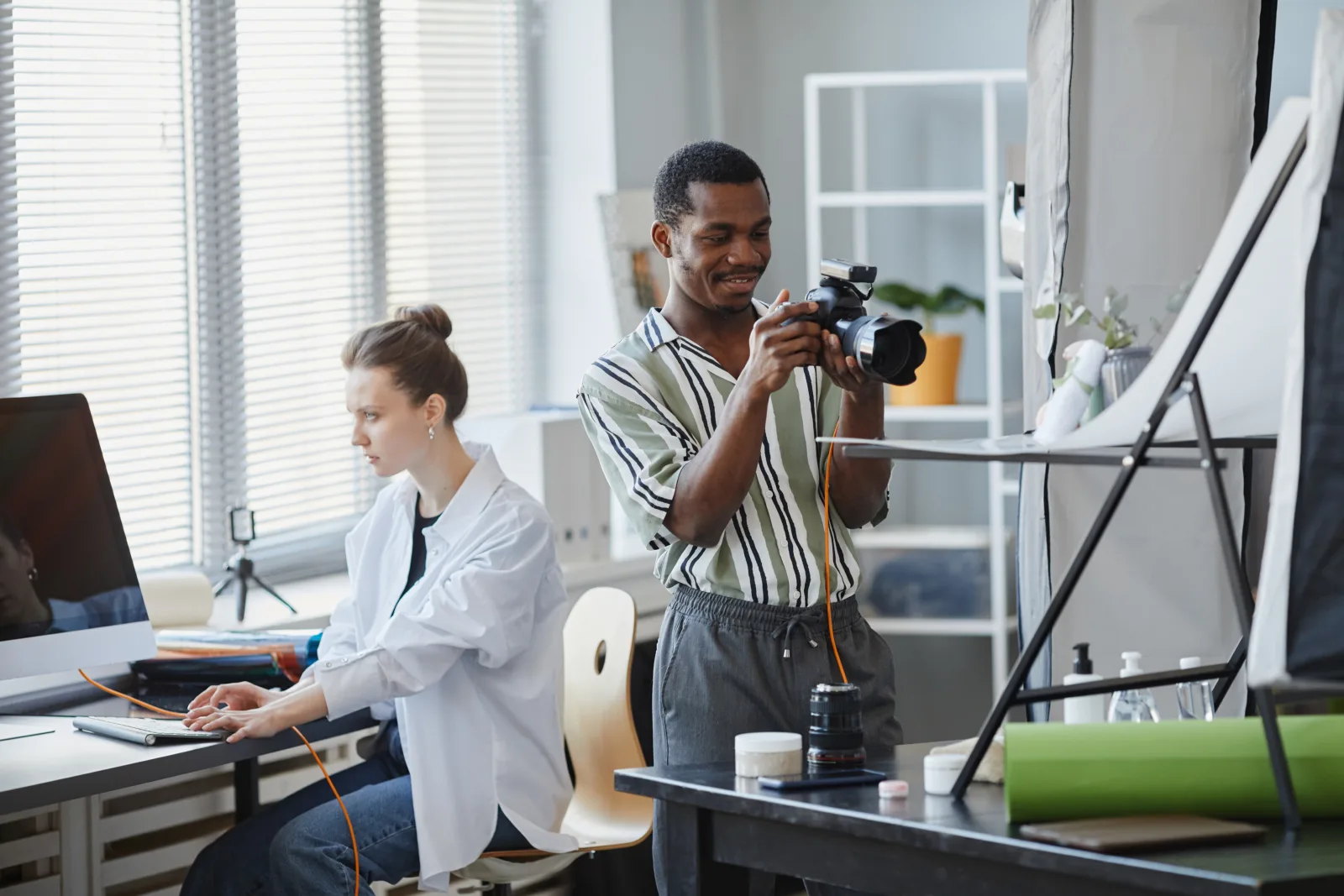 Ein Fotograf prüft seine Kamera in einem Studio, im Hintergrund arbeitet eine Kollegin am Schnittplatz