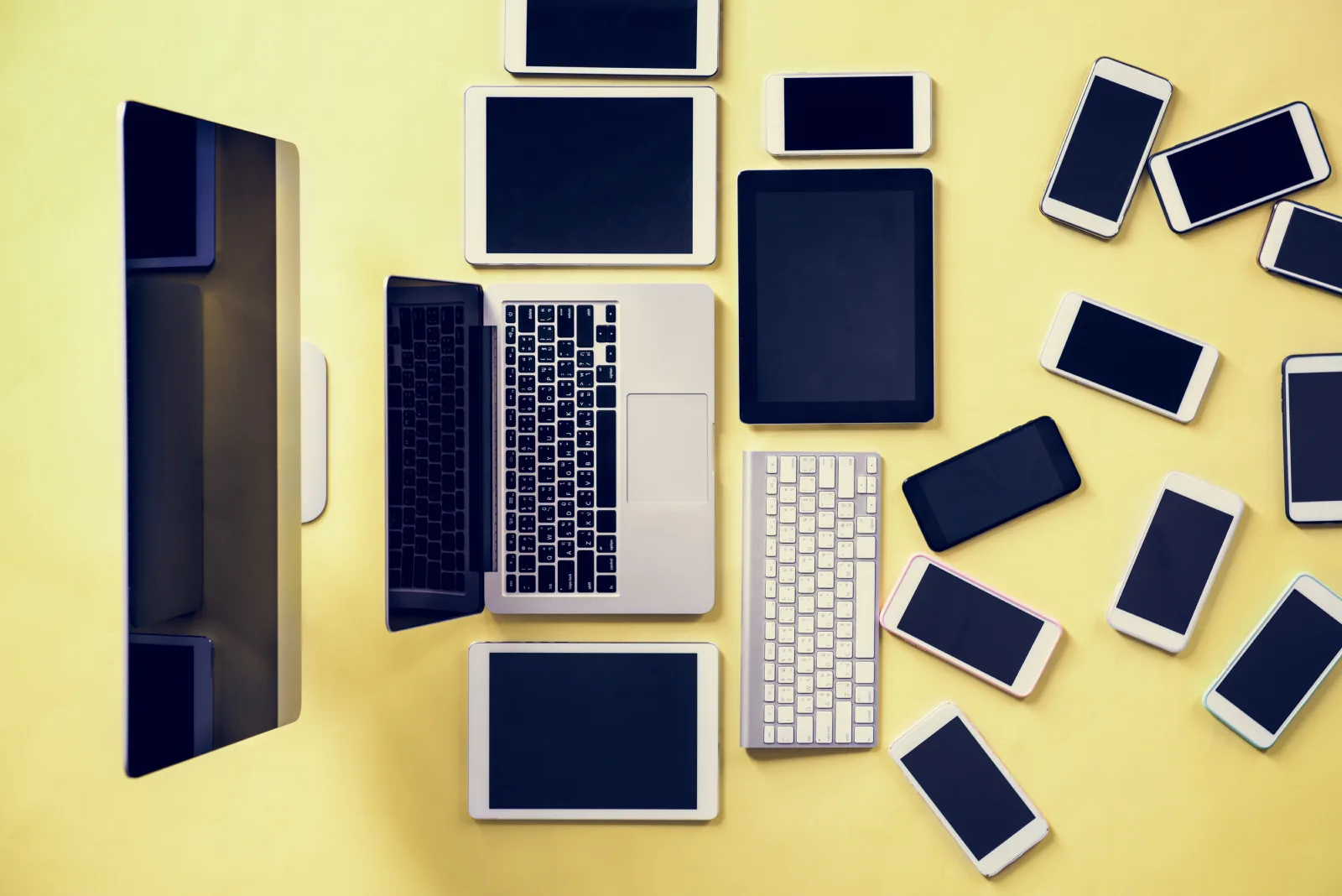 Bildschirm, Notebook, Tastatur, Tablets und mehrere Smartphones von oben auf gelbem Grund
