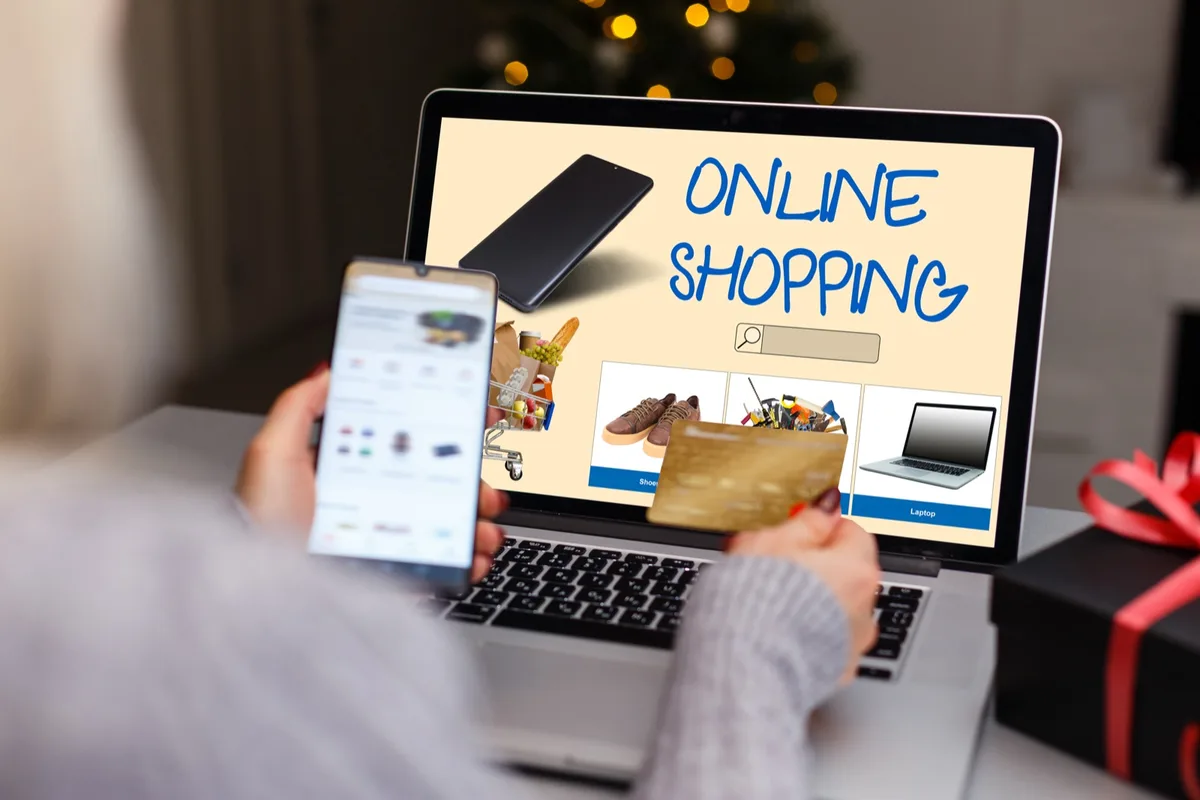 Eine Person hält ein Smartphone vor einen Laptop mit der Aufschrift Online Shopping, daneben ein Geschenk